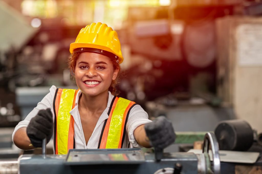 A young woman wearing a safety vest and hardhat smiles at the camera while operating a large machine.